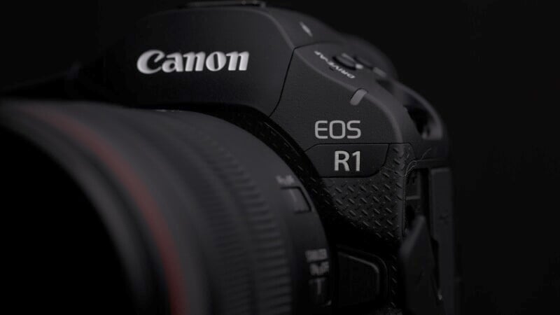 Close-up of a Canon EOS R1 camera. The image focuses on the camera's name and model details, featuring a textured black surface with subtle lighting revealing the design elements.