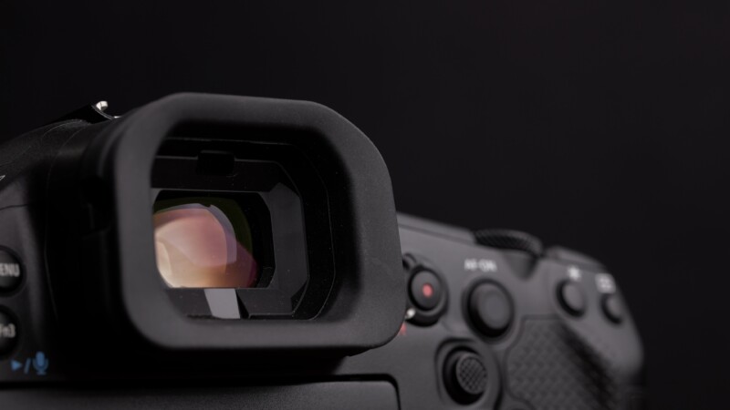 Close-up of a digital camera's viewfinder and control buttons against a dark background. The viewfinder is prominent, with light reflecting off the glass. The camera body appears detailed, showing various buttons and textured surfaces.