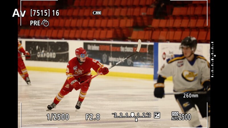 A hockey player in a red uniform skates on the ice, holding a stick with one hand. A player in a white uniform is in the foreground. The image edges display camera settings, including aperture, ISO, and shutter speed.