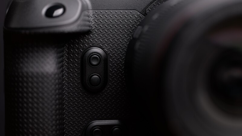 Close-up of a textured camera grip showing two circular buttons on the side of the camera body. The lens is partially visible on the right, blurred in the foreground.