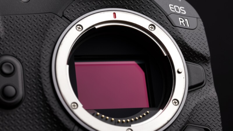 Close-up of a camera without a lens, showing the exposed sensor and mount. The camera body displays textured grips and the model marking "EOS R1" on the top right corner.