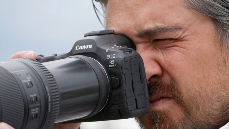 Close-up of a person taking a picture with a Canon EOS R5 camera. The person is looking through the camera's viewfinder, with one eye tightly closed. The camera has a large lens attached, and the background is blurred.