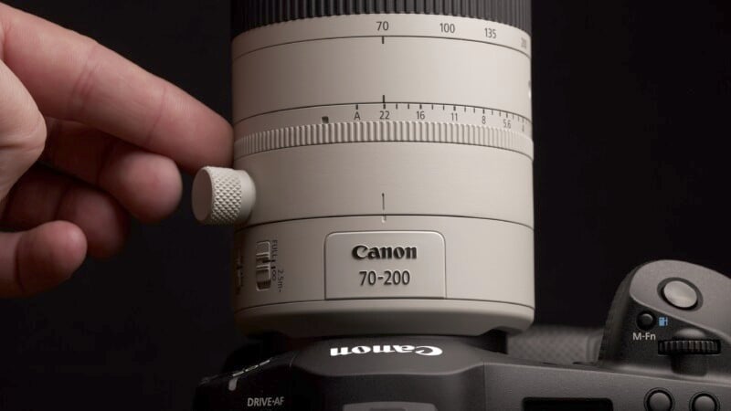 A close-up of a hand adjusting a white Canon 70-200mm camera lens mounted on a black Canon camera body. The focus is on the zoom ring and markings, indicating focal lengths from 70 to 200mm. The background is dark.