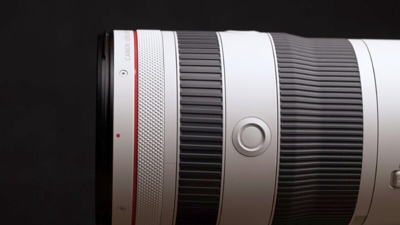 Close-up of a white camera lens with a black and red ring, featuring a textured zoom and focus ring. A small button is visible near the center, set against a black background.