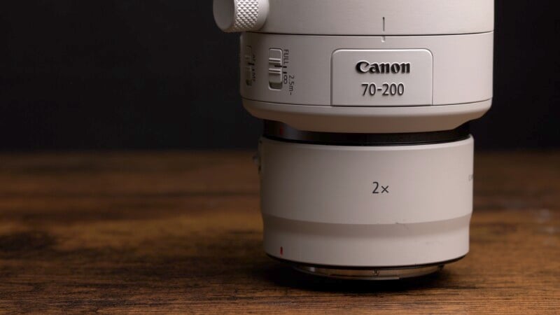 Close-up of a Canon 70-200mm camera lens with a 2x extender attached, placed on a wooden surface. The lens features switches and branding, and the background is dark, highlighting the equipment's details.