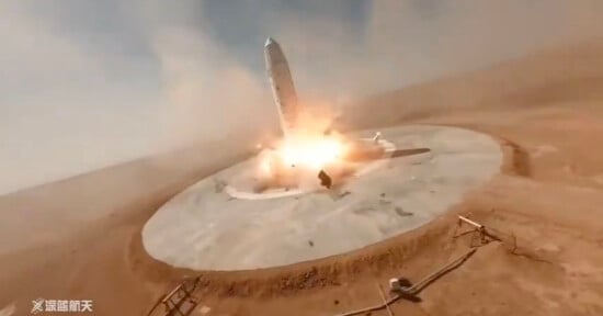 A rocket is landing vertically on a circular concrete pad in a desert. Flames and smoke are visible at the base of the rocket, indicating the engines are firing. The sky is clear, and the surrounding landscape is barren.