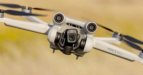 Close-up of a Mini 3 Pro drone in flight against a blurred background. The drone's camera and propellers are clearly visible, showcasing its design and features.