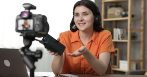 A woman in an orange shirt and headphones sits at a desk with a laptop and smartphone. She gestures towards a camera on a tripod, appearing engaged in a video recording or live streaming session. Shelves with books and decor are in the background.