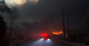 A fire truck with red lights illuminated drives down a smoke-filled street. Thick, dark smoke and flames are visible in the background, partially obscuring palm trees and a dimly lit sky. The scene appears intense and urgent.