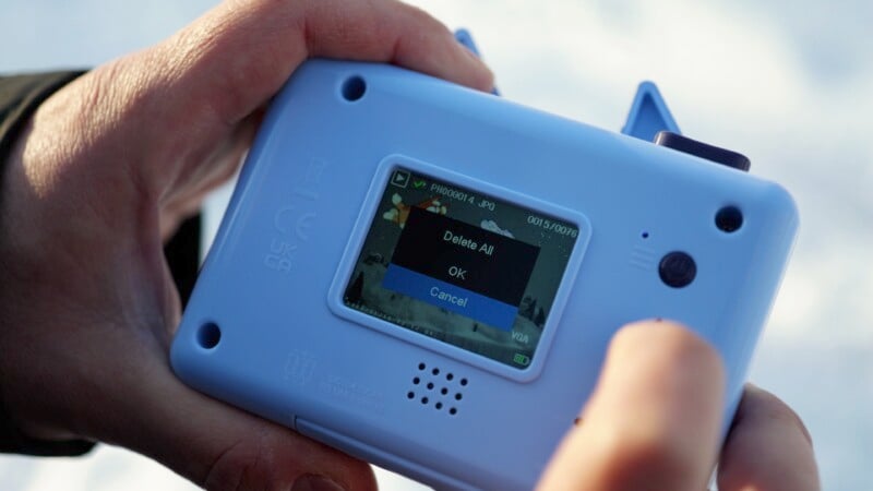 A person holds a small, blue digital camera with a screen displaying a prompt to delete all photos. Options shown are "Delete All," "OK," and "Cancel.