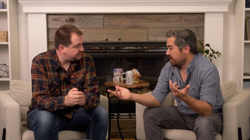 Two men are sitting on armchairs in front of a fireplace, engaged in a conversation. One man is wearing a plaid shirt and the other a denim shirt. A coffee table with decorative items and a can is visible between them. They appear animated and focused.