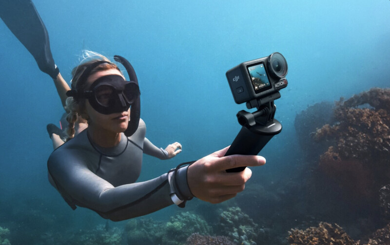 A person wearing a wetsuit and snorkeling gear swims underwater, holding a camera on a handle. Coral and marine life are visible in the background, with blue ocean water surrounding them.