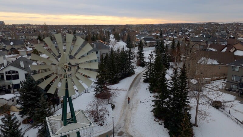 A large windmill in the foreground overlooks a snowy suburban neighborhood at sunset. A person walks along a snow-covered path lined with tall evergreen trees. Houses extend into the distance under a cloudy sky.