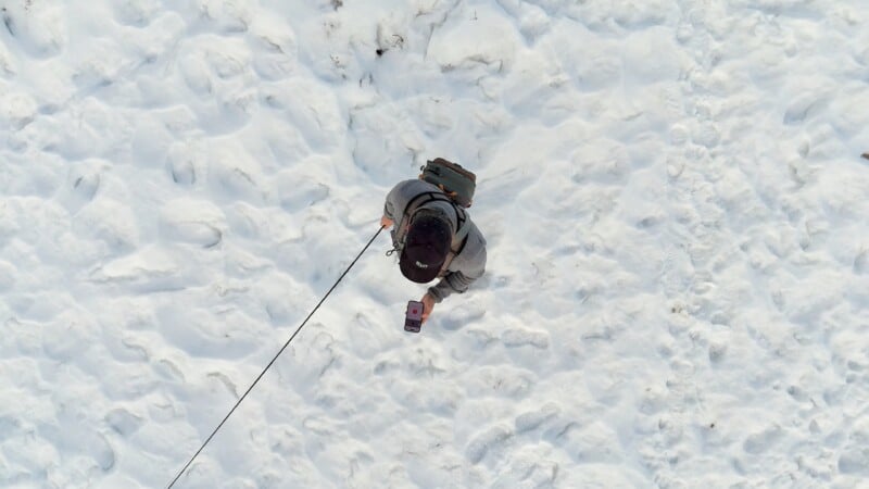 A person walking on a snow-covered ground viewed from above. The person is holding a leash with a small animal, possibly a dog, and a phone. They're wearing a dark hat and a backpack. The snow is uneven with visible footprints.