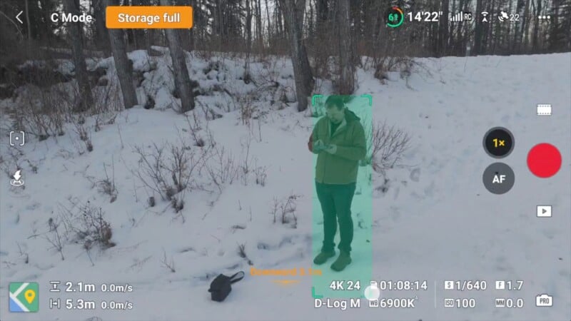 A person stands in a snowy forest, looking down at a device in their hands. Snow covers the ground and trees. Camera interface details are visible, indicating a full storage warning and settings information.