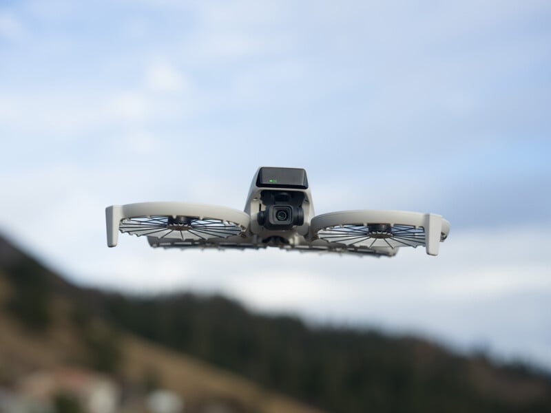 A drone equipped with a camera hovers mid-air against a backdrop of a cloudy sky and distant mountains. Its design includes protective guards around the propellers.