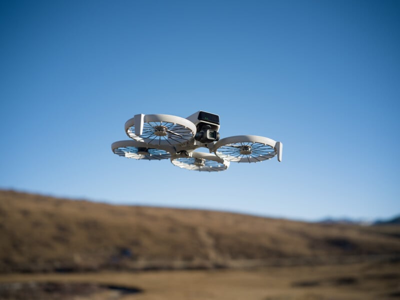 A white drone with a sleek design is flying over a dry, grassy landscape. The sky is clear blue, and the distant mountains are visible on the horizon.