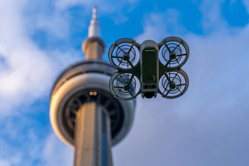 A drone hovers in front of a tall tower, capturing a skyward view. The sky is cloudy, and the tower stretches above, creating a striking urban scene against the backdrop.