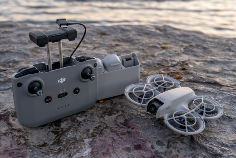 A grey DJI drone with protective guards rests on a rocky surface near a body of water. Beside it is a matching remote controller with a screen holder and visible control sticks. The water's surface is calm with a gentle reflection of the sky.