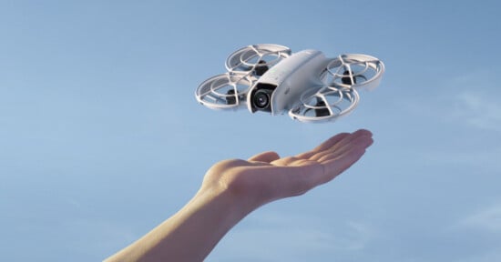 A person's hand is reaching up as a small, white drone with four enclosed propellers hovers above it against a clear blue sky. The drone appears to be equipped with a camera on its front.