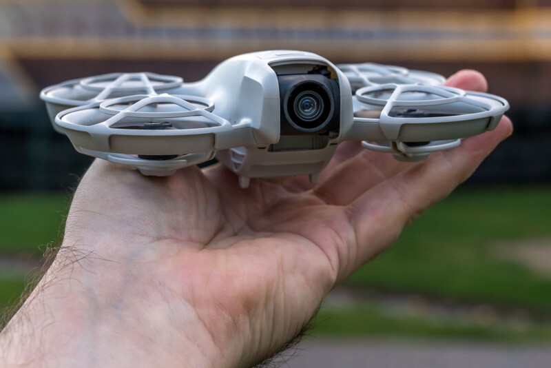 A person holds a small, white quadcopter drone with protective guards around its propellers, outdoors. The drone's camera is visible, and the background includes blurred greenery and a building.