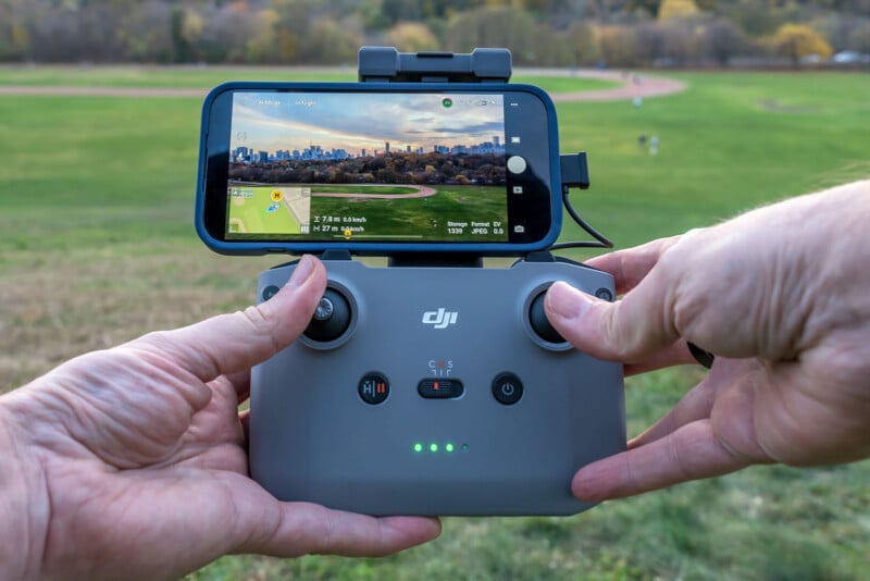 A person holds a DJI drone remote controller with a smartphone attached. The smartphone displays a live video feed of a green field and distant city skyline. The surroundings include grass and trees under a clear sky.