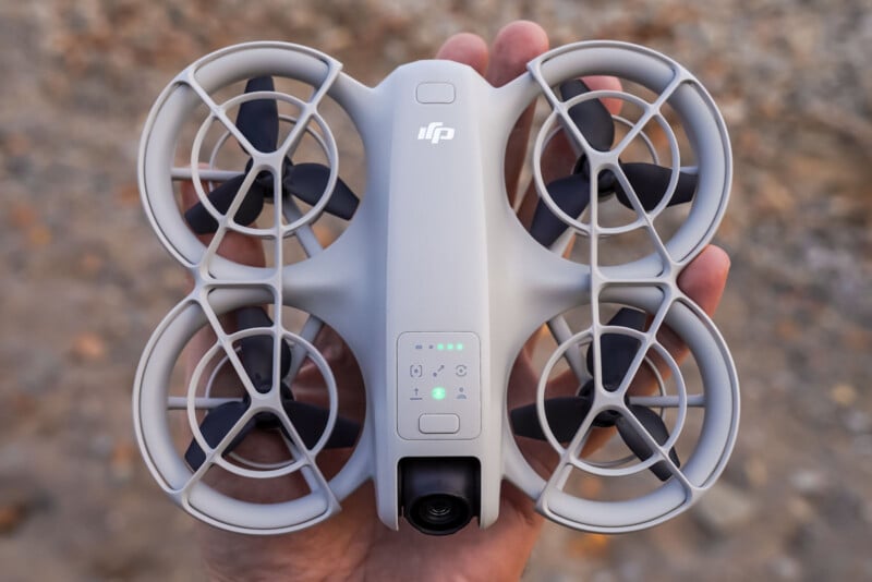 A hand holds a small, gray drone with four circular propeller guards. The drone has a logo on top and an LED indicator light. The background is a blurred outdoor setting.