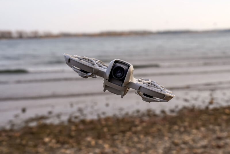 A small drone with a camera hovers over a rocky beach, with the sea and a cloudy sky in the background.