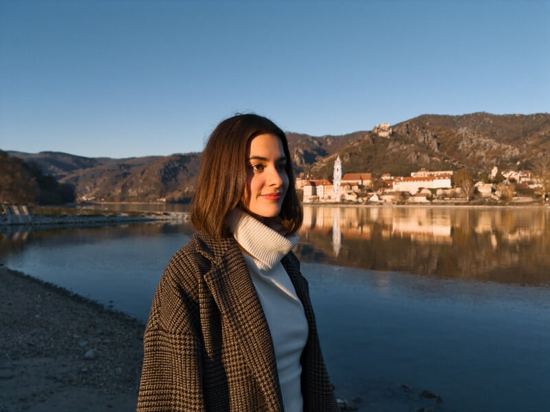 A woman with shoulder-length hair and a checkered coat stands by a calm river. The background features a picturesque town with rustic buildings and hilly terrain under a clear blue sky.