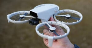 A person holding a compact white drone with four circular propellers. The drone features a camera mounted at the front. The background is blurred, emphasizing the drone's details.