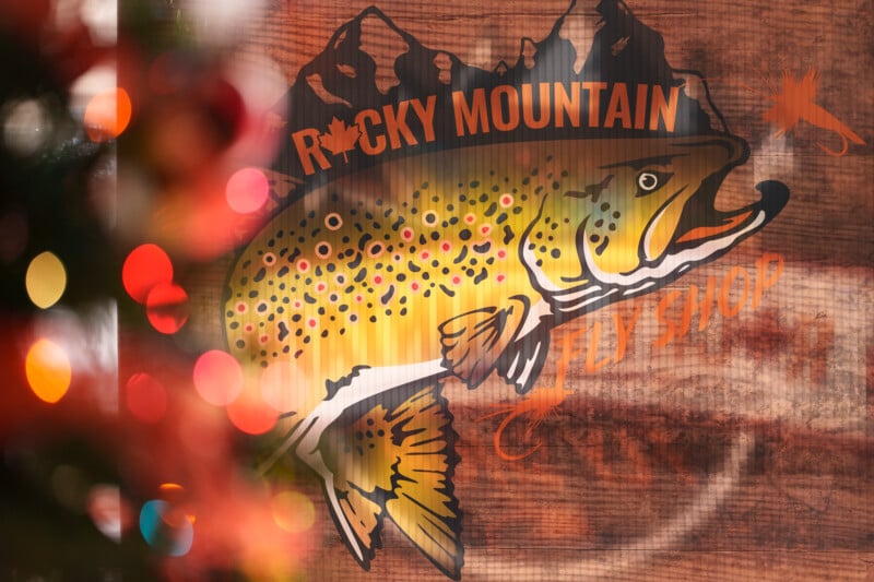 A festive scene features a blurred Christmas tree on the left, with colorful lights. To the right is a painted sign of a trout fish and text reading "Rocky Mountain Fly Shop" against a wooden background.