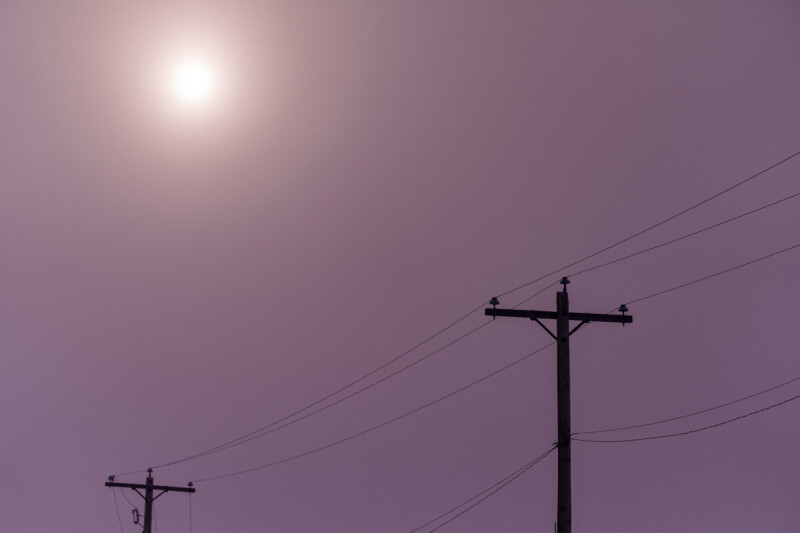 Two silhouettes of power lines and poles against a hazy, purple-tinted sky with a faint sun in the background, creating an ethereal and minimalistic scene.