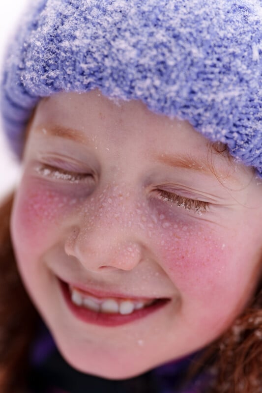 A close-up of a smiling child with eyes closed, wearing a blue knitted hat covered in snowflakes. The child's cheeks are rosy, and there are tiny droplets of water on their eyelashes and skin.