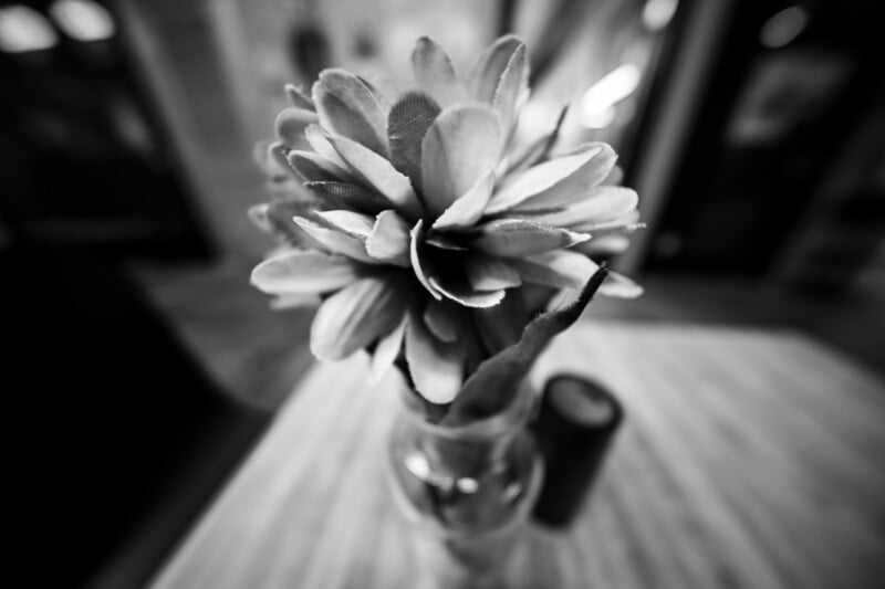 A black and white close-up of a flower in a glass vase on a wooden table. The focus is on the detailed petals, with a blurred background providing a soft, artistic effect.