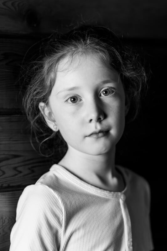 Black and white portrait of a young girl with light hair, wearing a light-colored top. She's standing against a wooden wall, looking directly at the camera with a neutral expression. The lighting highlights her facial features.