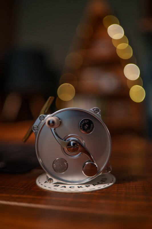 A close-up image of a vintage fishing reel placed on a round coaster with "BACKPACKER" written on it. The background is softly blurred, featuring warm-toned bokeh lights and indistinct shapes.