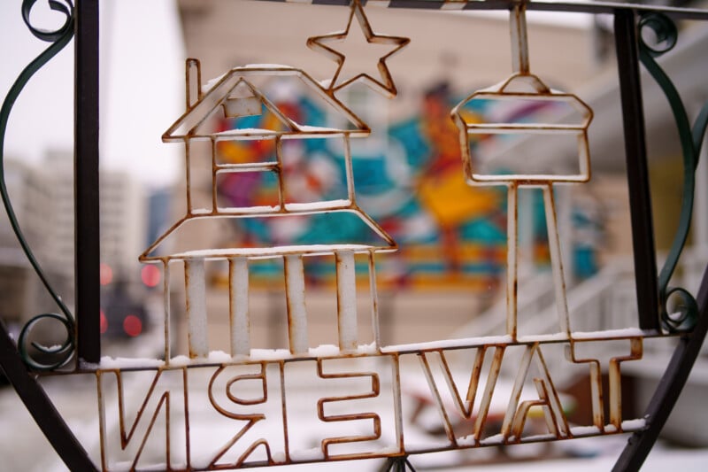 Close-up of a rusted metal gate adorned with a house, TV tower, and a star. Snow lightly covers parts of the design. A colorful, blurred mural is visible in the background.