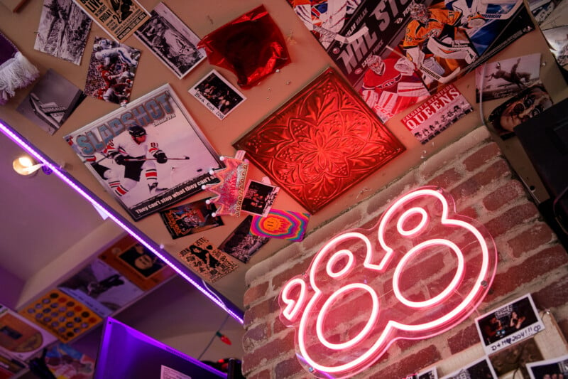 A vibrant wall collage features sports posters, art, and memorabilia under warm lighting. A neon pink '88 sign stands out against a brick surface, adding a retro touch to the eclectic array of images and textures.