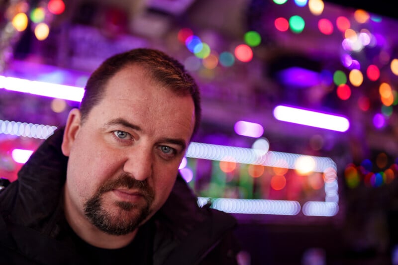 A man with a beard and a serious expression stands in a vibrant setting filled with colorful, blurred lights in the background. He wears a dark jacket, and the atmosphere appears lively and festive.