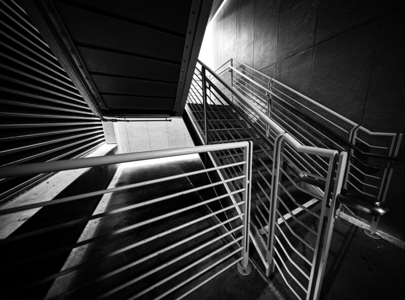 Black and white image of a modern industrial staircase with metal railings. The light casts sharp geometric shadows on the concrete floor, creating an abstract pattern. The perspective emphasizes the angles and lines of the structure.