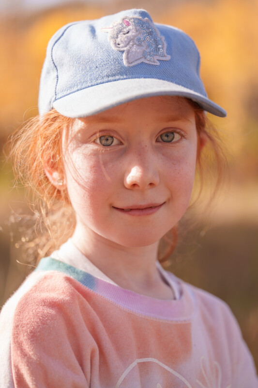 A young girl with red hair wearing a blue cap with a unicorn design and a pink sweater stands outdoors. She is looking at the camera with a calm expression. The background is blurred, featuring autumn colors.