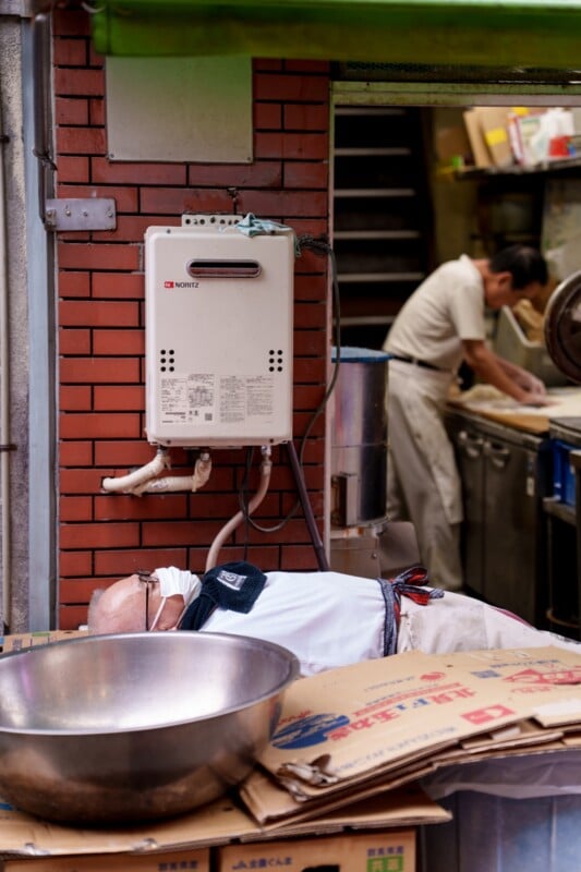 A person is sleeping on a stack of cardboard boxes next to a brick wall, beneath a wall-mounted appliance. Another person is busy working in a kitchen in the background. A large metal bowl is in the foreground.