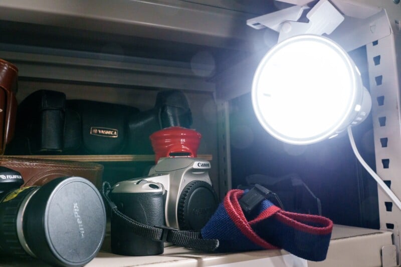 A shelf displaying a vintage Canon camera with a strap, a Fujifilm lens cap, a red jar, and leather camera cases. A bright circular light is mounted above, illuminating the scene.