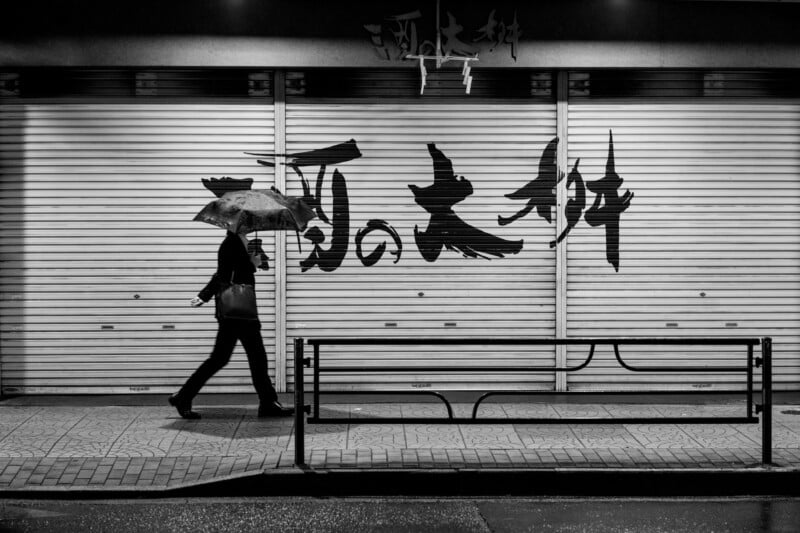 A person walks with an umbrella in front of a closed shop with large, bold Japanese calligraphy on the shutters. The image is in black and white, highlighting the contrast and texture of the scene.