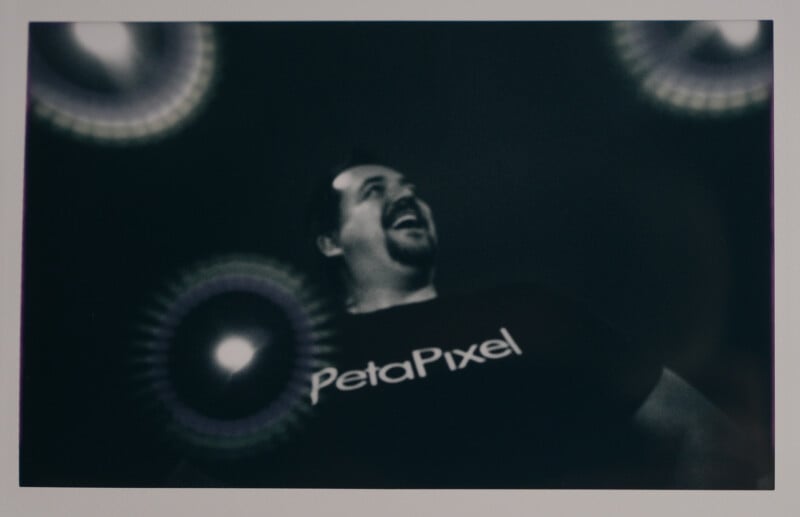 A man laughing, wearing a "PetaPixel" t-shirt, is captured in black and white. Light reflections appear around him against a dark background, adding an abstract element to the image.