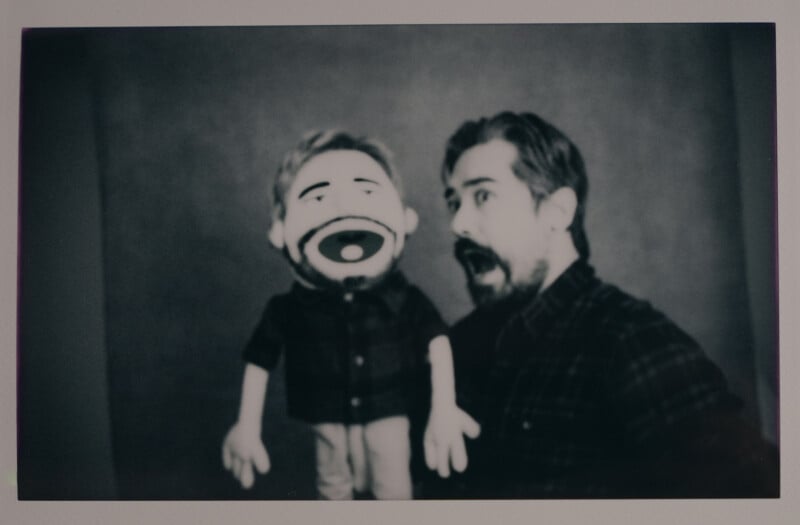 A person with facial hair and a plaid shirt is posing excitedly next to a puppet wearing a similar outfit. Both have wide open mouths, mimicking an expression of surprise or excitement. The background is plain and blurred.