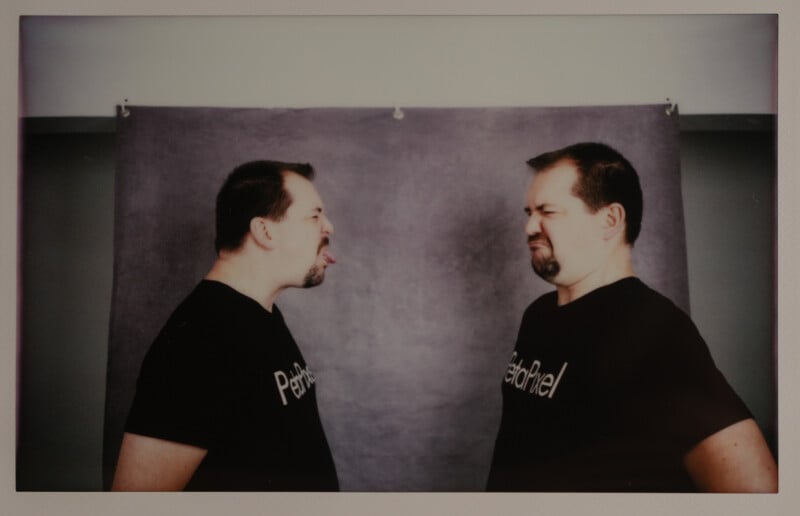 Two men with similar features wearing black T-shirts stand facing each other. One man sticks out his tongue playfully while the other makes a funny, displeased expression. A gray backdrop is behind them.