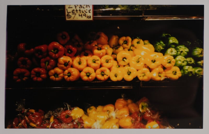 A grocery display with rows of red, yellow, and green bell peppers. A sign above reads "2 pack lettuce $7.49 ea." The peppers are stacked neatly in vibrant colors under the store lights.