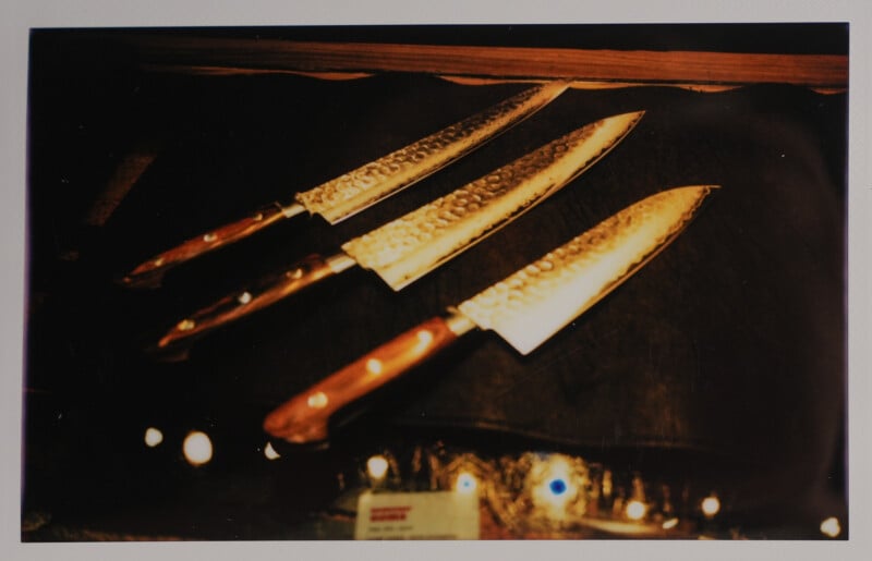 A set of three kitchen knives with hammered steel blades and wooden handles are displayed on a dark surface. The blades catch the light, creating a shiny effect. Small lights or reflections are visible at the bottom of the image.