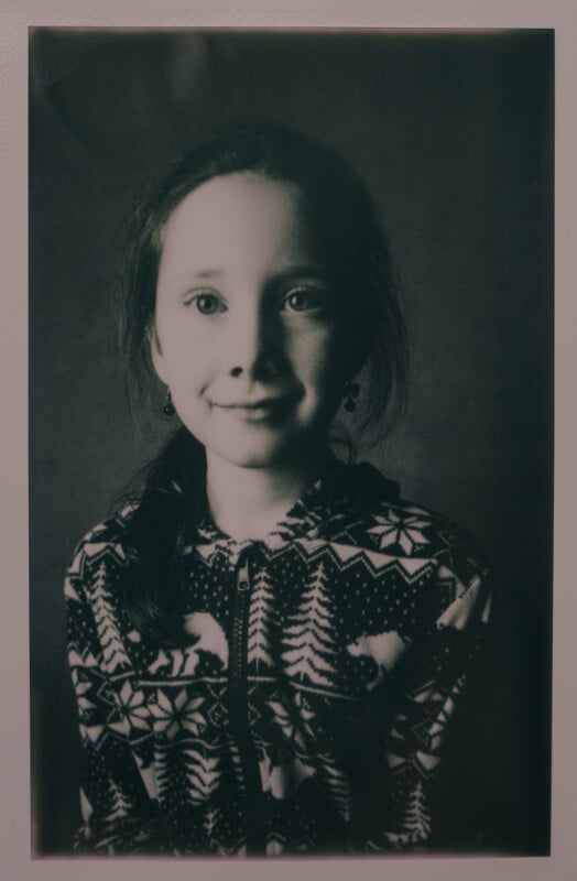 Black and white portrait of a young girl with a serene expression, wearing a patterned sweater with trees and reindeer. She has long hair tied back, and the background is dark and plain.